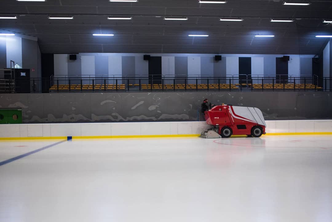 Montovaná ocelová oblouková hokejová hala Bruck – tribuny s žlutými sedadly a rolba upravující ledovou plochu, moderní interiér s akustickými panely, realizace HUPRO SYSTEMS SE.