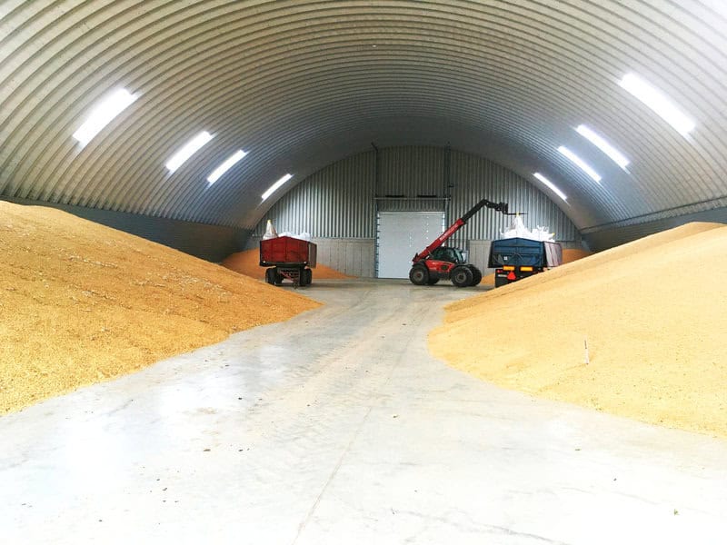 Interiér montované ocelové obloukové zemědělské haly využívané ke skladování obilí, se zemědělskou technikou a hromadami zrnin, realizace HUPRO SYSTEMS SE.