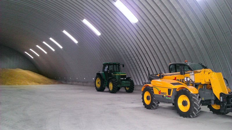Interiér ocelové obloukové zemědělské haly se zemědělskou technikou značek John Deere a Dieci a uskladněným obilím v pozadí. Realizace HUPRO SYSTEMS SE.