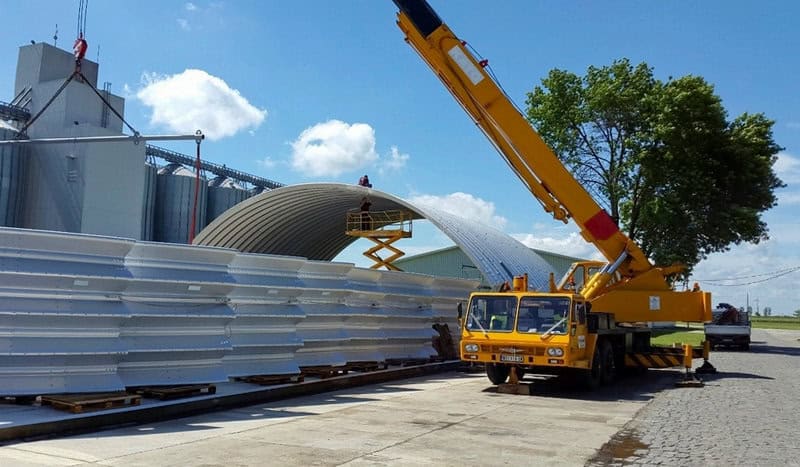 Montáž ocelové obloukové haly pomocí jeřábu a pracovní plošiny, s díly konstrukce připravenými na paletách. Realizace HUPRO SYSTEMS SE.