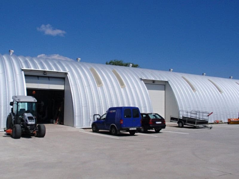 Ocelová oblouková hala s několika vraty, zaparkovanými vozidly a zemědělskou technikou před vchodem. Realizace HUPRO SYSTEMS SE.