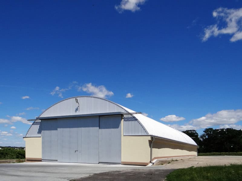 Ocelová oblouková montovaná zemědělská hala se světlou fasádou a velkými posuvnými vraty, vhodná pro skladování techniky či úrody. Realizace HUPRO SYSTEMS SE.