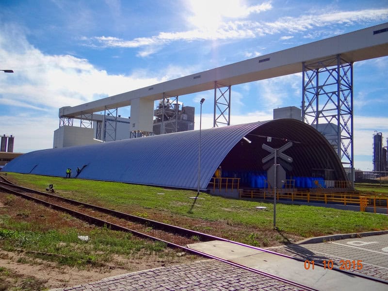 Velká průmyslová ocelová oblouková hala umístěná u železniční tratě, využívaná pro skladování a manipulaci s materiálem.