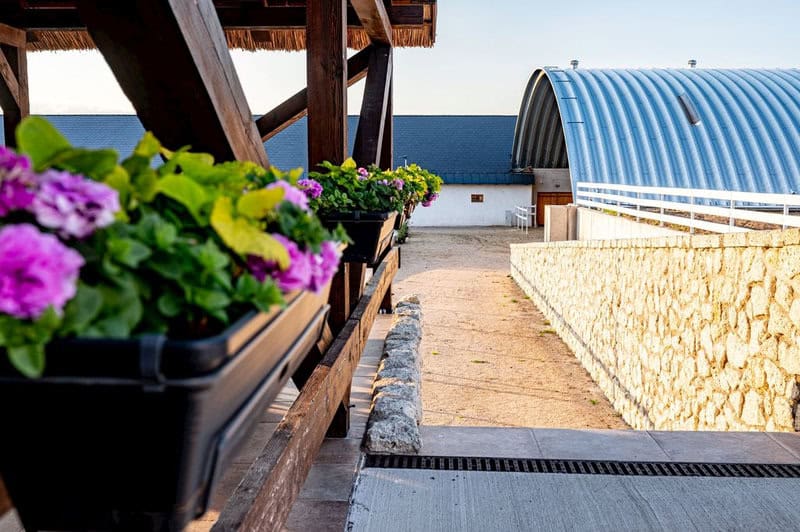 Moderní modrá oblouková zemědělská hala HUPRO SYSTEMS SE vedle kamenné zdi a květinové terasy ve venkovském areálu.