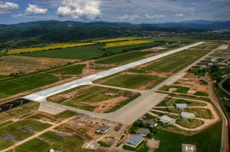 Obloukové ocelové montované armádní haly na vojenském letišti obklopeném zelenou krajinou a horami, využívané pro údržbu a parkování letounů, samonosná konstrukce HUPRO SYSTEMS SE