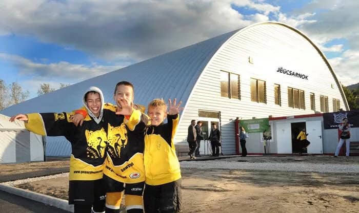 Moderní hokejová hala Budapešť s obloukovou ocelovou konstrukcí, před kterou pózují mladí hokejisté v klubových dresech, realizace HUPRO SYSTEMS SE.