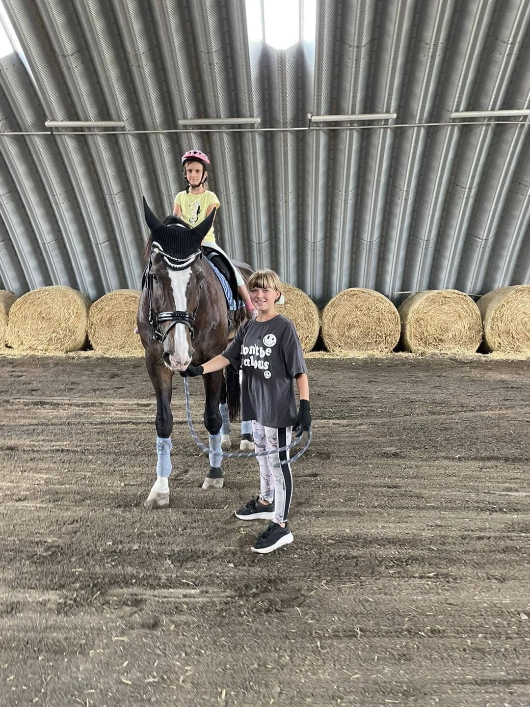 Dívka vede koně s jezdkyní uvnitř jízdárny v Břeclavi s obloukovou ocelovou konstrukcí a balíky sena v pozadí. Realizace HUPRO SYSTEMS SE.