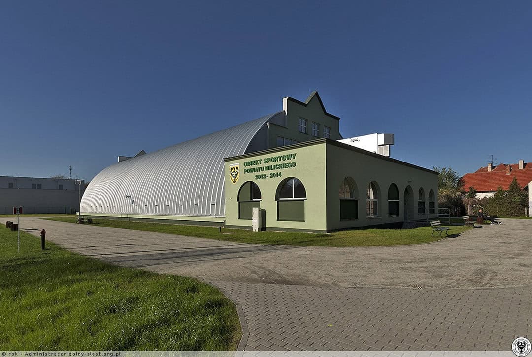 Montovaná ocelová oblouková víceúčelová sportovní hala Milicz, exteriér se vstupní částí a nápisem Obiekt Sportowy Powiatu Milickiego. Realizovala společnost HUPRO SYSTEMS SE.