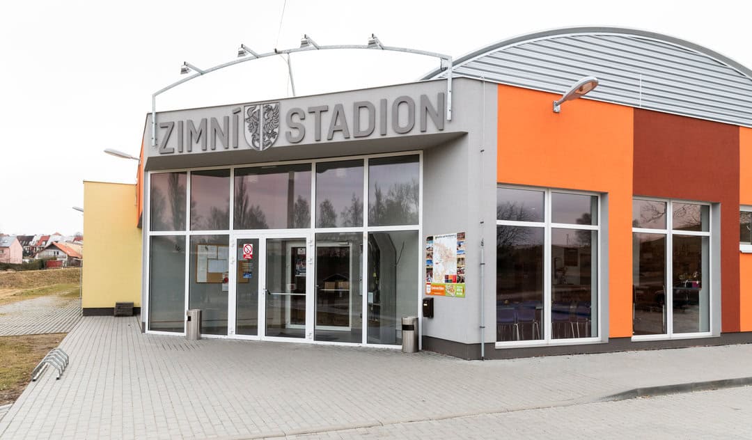 Vstupní část zimního stadionu v Bystřici nad Pernštejnem s nápisem „Zimní stadion“, moderní fasádou v oranžových odstínech a proskleným vstupem. Realizace HUPRO SYSTEMS SE.