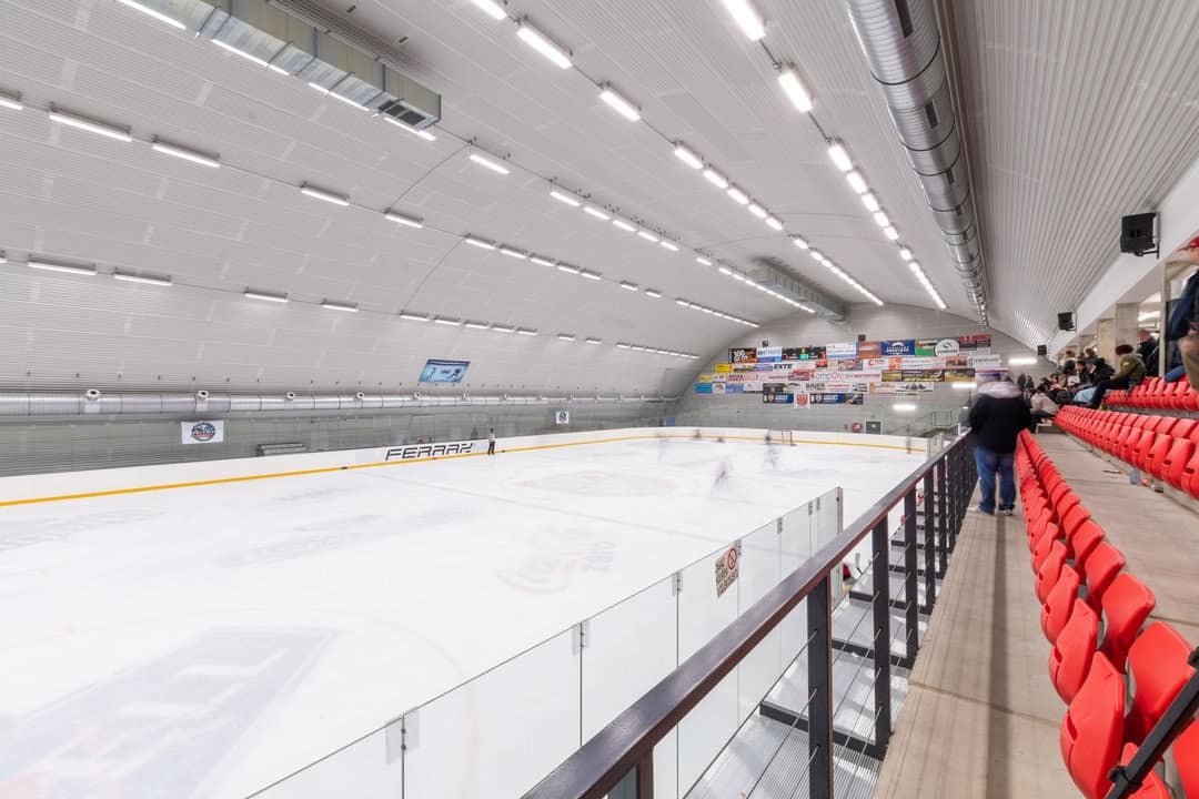 Zimní stadion Telč – interiér moderní obloukové montované ocelové haly s ledovou plochou, tribunou s červenými sedadly a hokejovým zápasem, realizace HUPRO SYSTEMS SE.