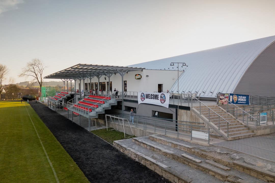 Zimní stadion Telč – pohled na tribunu s červenými sedadly a vstupní část moderní obloukové montované ocelové haly od firmy HUPRO SYSTEMS SE.