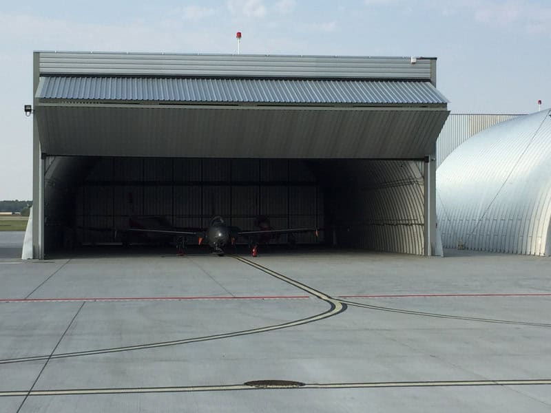 Obloukové ocelové montované letecké haly s otevřenými vraty a uskladněnými letadly na letištní ploše. Realizace HUPRO SYSTEMS SE.