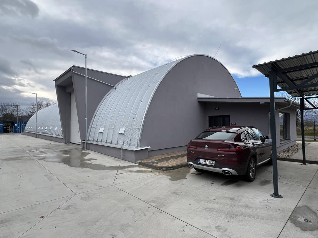 Exteriér soukromého muzea automobilů v obloukové montované ocelové hale od HUPRO SYSTEMS SE, s moderní šedou fasádou a přilehlým parkovištěm.