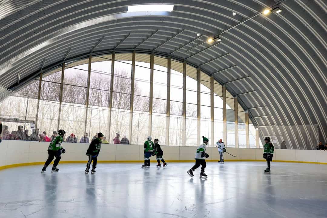 Dětský hokejový trénink v obloukové montované ocelové hale ve městě Hurbanovo na Slovensku. Krytá ledová plocha s prosklenou čelní stěnou a moderním osvětlením.