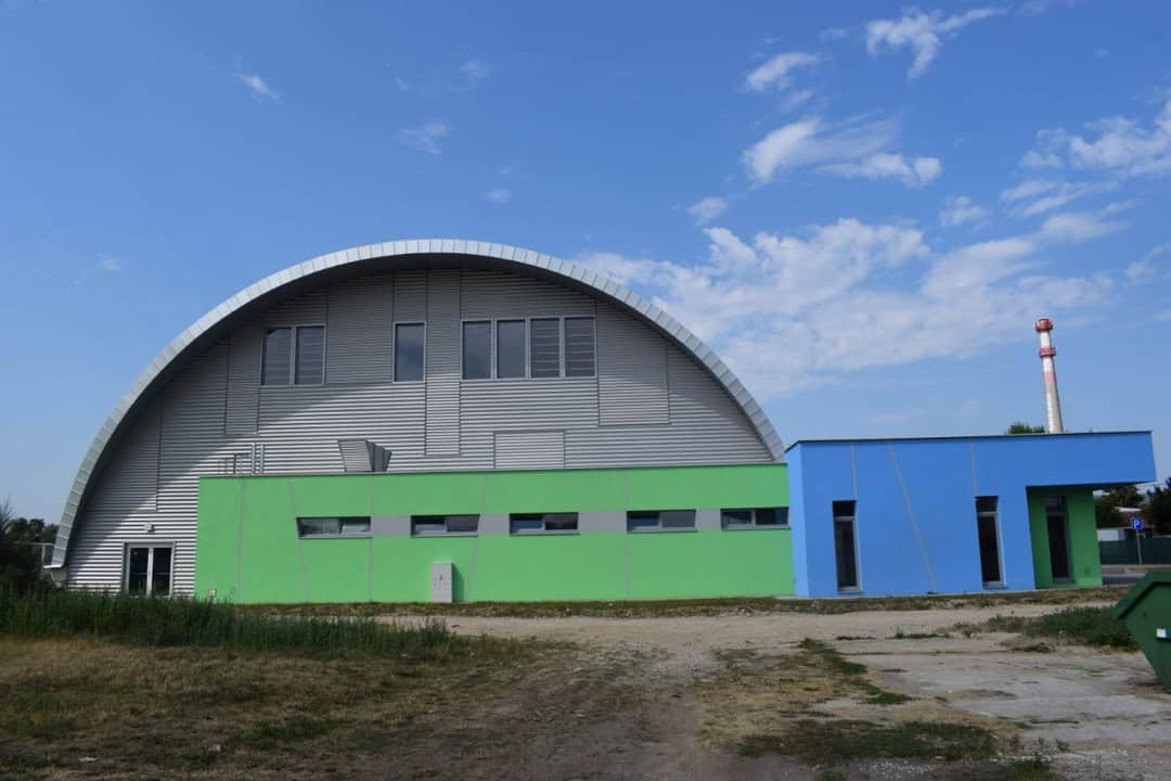 Exteriér moderní sportovní haly s obloukovou střechou ve slovenském městě Bytča. Budova má kombinaci šedé, zelené a modré fasády a je dílem společnosti HUPRO SYSTEMS SE.