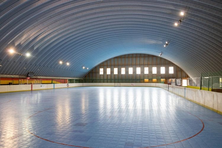 Pohled přes modrou hrací plochu v obloukové sportovní hale v Bratislavě Ružinově, postavené systémem HUPRO SYSTEMS SE, s výrazným klenutým stropem a řadou oken v zadní stěně.