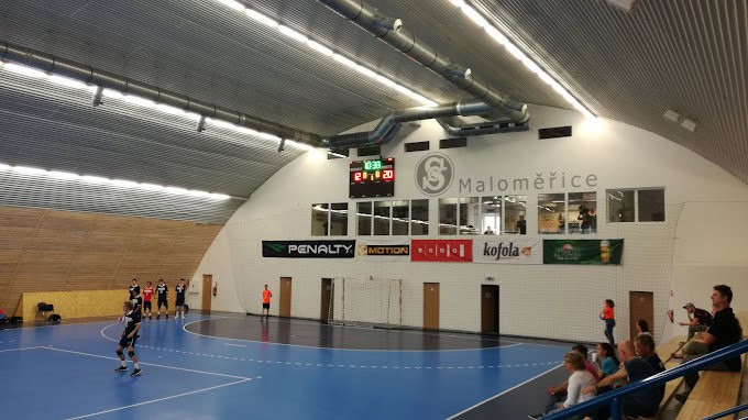 Zápas házené ve víceúčelové sportovní hale v Brně-Maloměřicích s tribunou a logem Maloměřice na stěně.