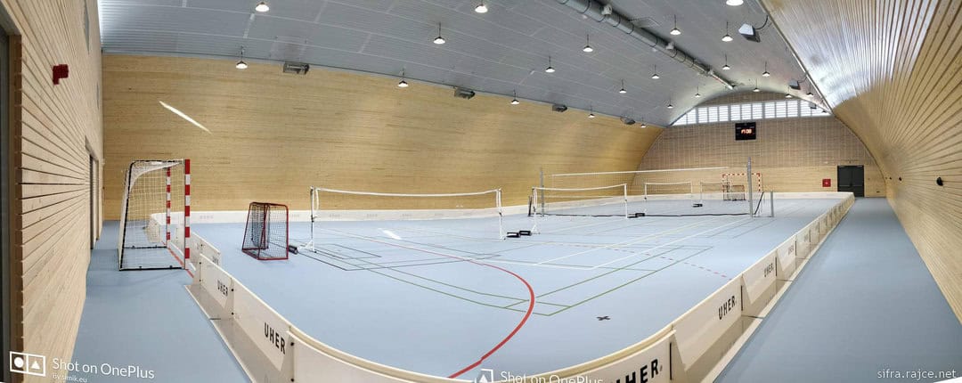Panoramatický pohled na interiér sportovní haly v Hodonicích s modrým povrchem, dřevěnými stěnami a hřištěm pro různé sporty, halu realizovala společnost HUPRO SYSTEMS SE.