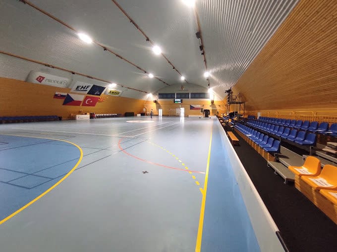 Interiér ocelové sportovní haly v Kynžvartu s multifunkčním hřištěm, tribunou a vlajkami nad hrací plochou, realizace HUPRO SYSTEMS SE.