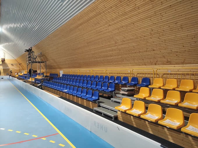 Tribuna s modrými a žlutými sedačkami v ocelové sportovní hale v Kynžvartu s dřevěným obložením stěn, realizace HUPRO SYSTEMS SE.