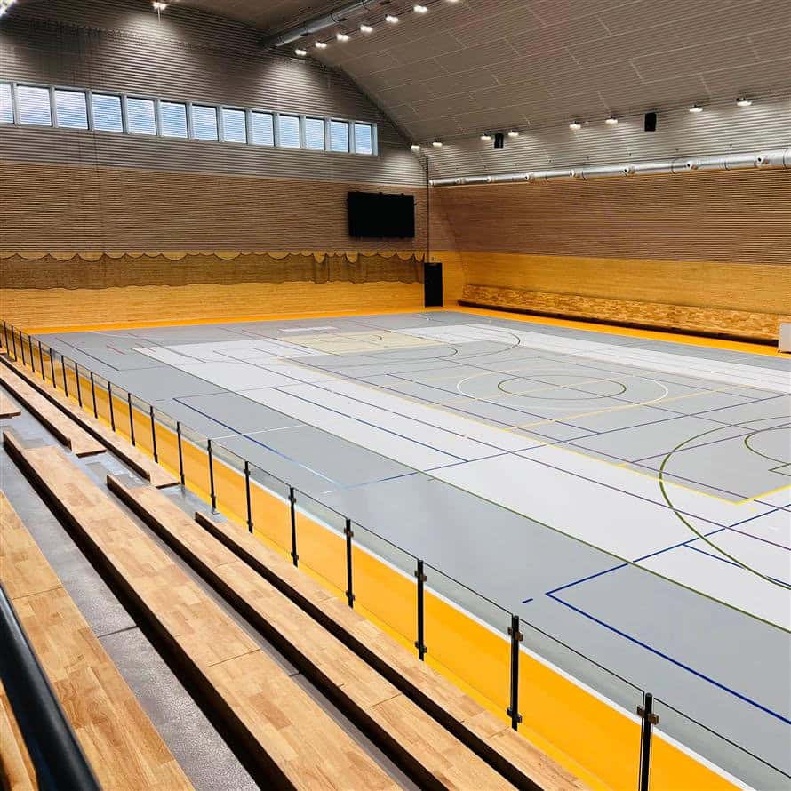 Moderní interiér obloukové montované ocelové sportovní haly Háje ve Slaném s multifunkčním hřištěm, dřevěnými tribunami a LED osvětlením. Stavba roku 2024.