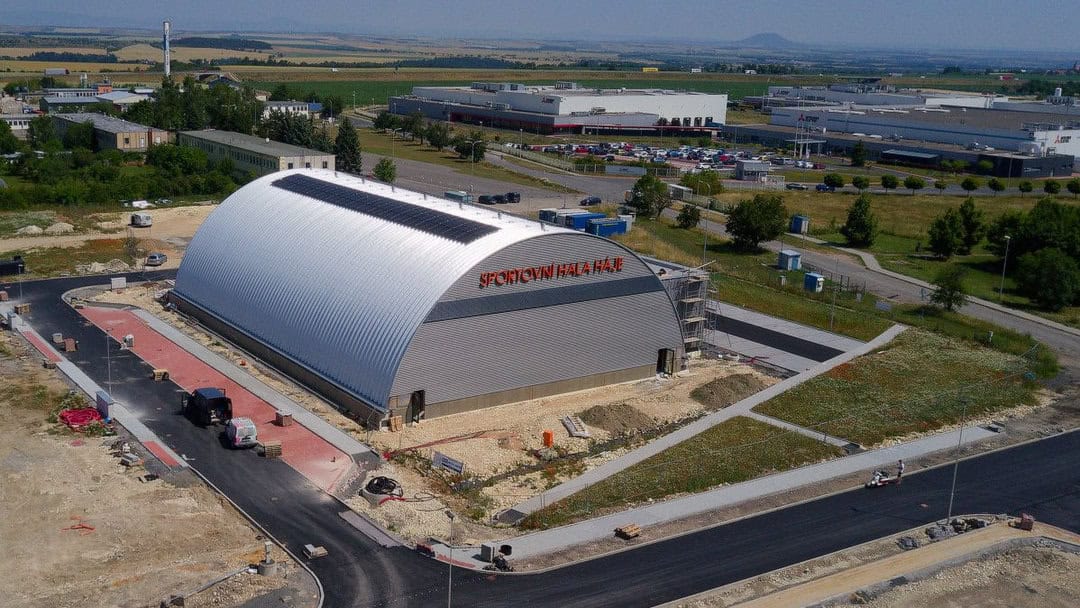 Letecký pohled na obloukovou montovanou ocelovou sportovní halu Háje ve městě Slaný během dokončovacích prací. Moderní víceúčelová hala oceněná titulem Stavba roku 2024.