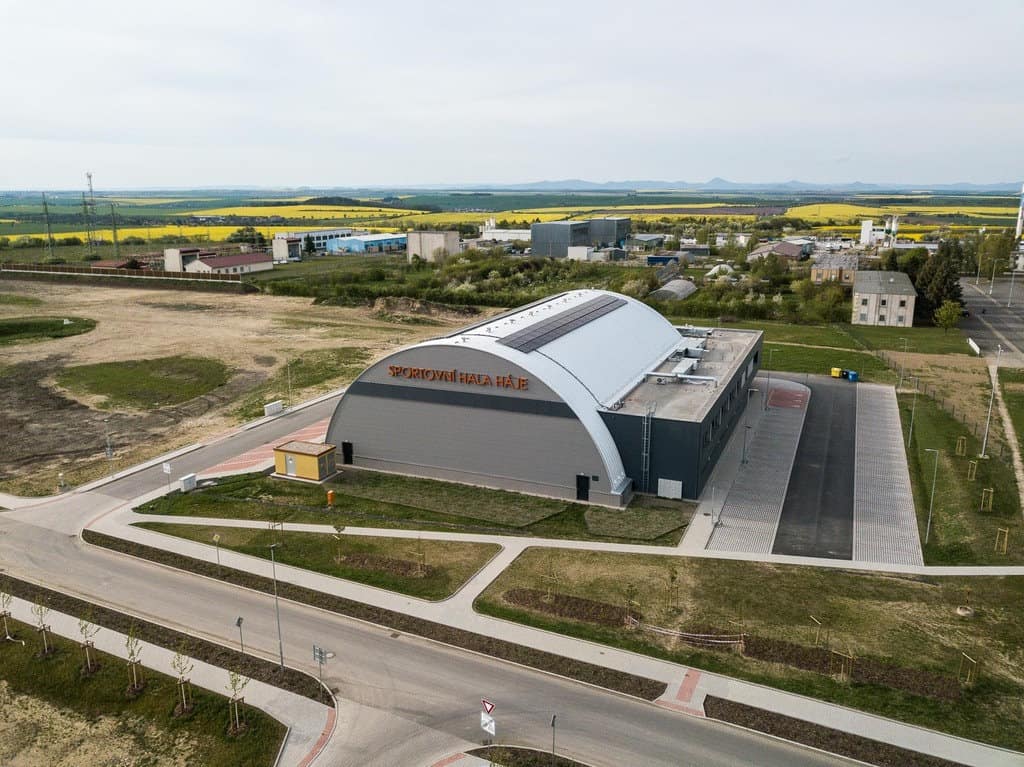 Letecký snímek moderní obloukové montované ocelové sportovní haly Háje ve městě Slaný, oceněné jako Stavba roku 2024. Energeticky úsporná víceúčelová budova s fotovoltaickými panely na střeše.