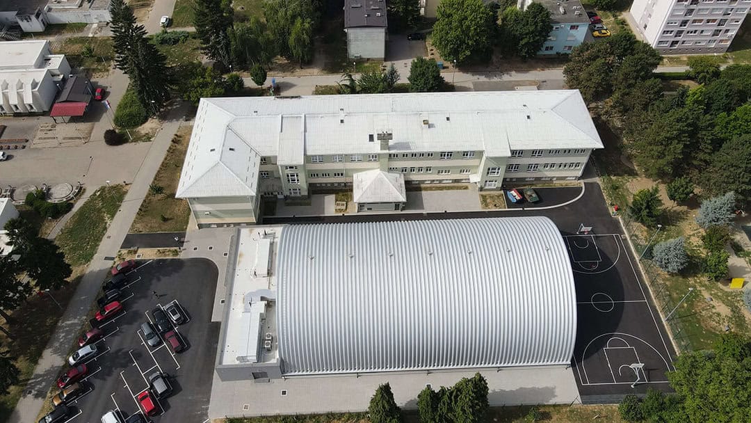 Víceúčelová montovaná ocelová sportovní hala Koprivnica, letecký pohled na moderní sportovní halu HUPRO SYSTEMS SE připojenou ke školní budově s venkovním hřištěm.