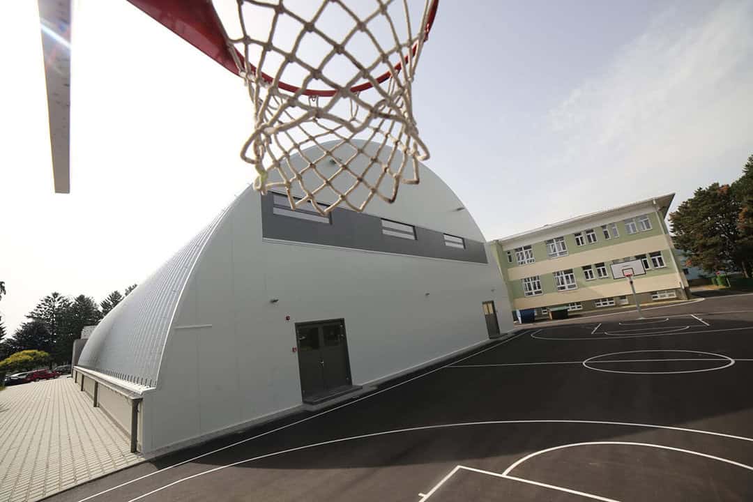 Víceúčelová montovaná ocelová sportovní hala Koprivnica s venkovním basketbalovým hřištěm, moderní sportovní areál HUPRO SYSTEMS SE u školní budovy.