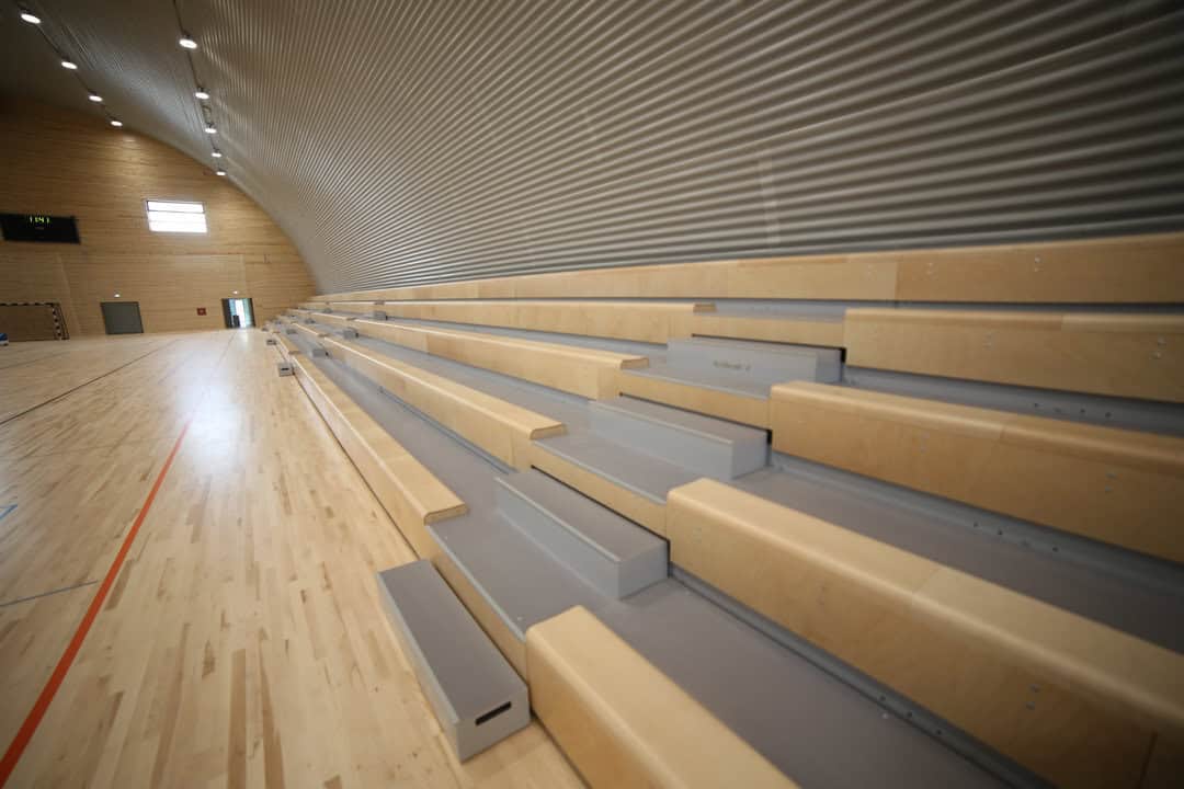 Oblouková sportovní hala Křiževci, interiér s dřevěnou sportovní podlahou a tribunou pro diváky z pohledu podél hrací plochy, realizace HUPRO SYSTEMS SE.