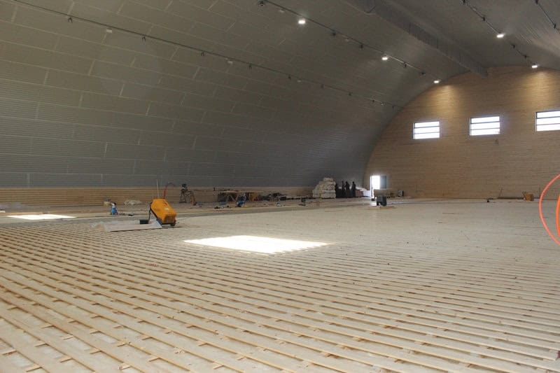 Oblouková sportovní hala Křiževci ve fázi montáže, interiér s rozpracovanou konstrukcí sportovní podlahy a dřevěným obložením čelní stěny, realizace HUPRO SYSTEMS SE.