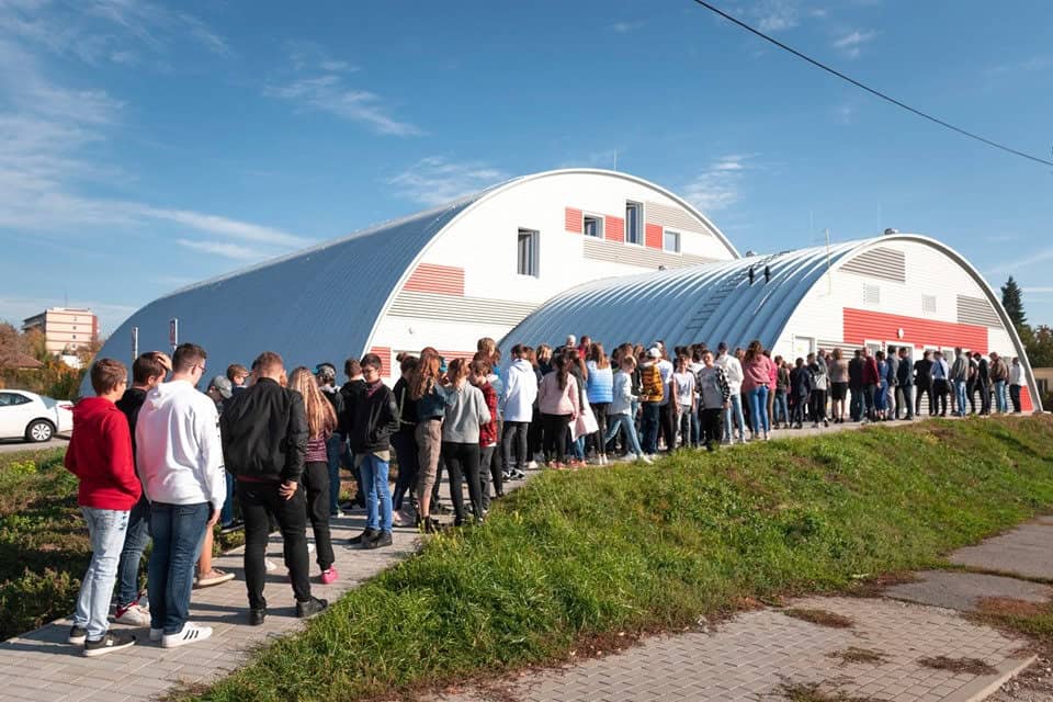 Sportovní hala Nitra Tulipanová – moderní oblouková stavba HUPRO SYSTEMS SE s červeno-šedou fasádou, před kterou stojí skupina studentů při slavnostním otevření.