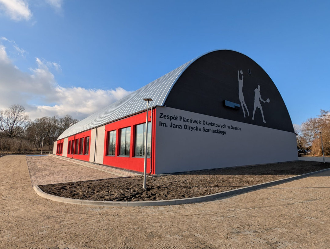 Montowana stalowa łukowa hala sportowa Szaniec, elewacja zewnętrzna w kolorze czerwono-szarym i strefa wejściowa. Realizacja firmy HUPRO SYSTEMS SE. Montowana stalowa łukowa hala sportowa Szaniec, elewacja zewnętrzna w kolorze czerwono-szarym i strefa wejściowa. Realizacja firmy HUPRO SYSTEMS SE.