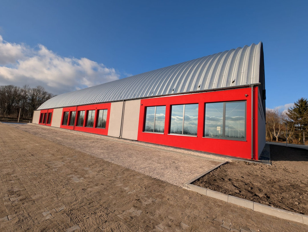 Widok boczny montowanej stalowej łukowej hali sportowej Szaniec z przeszkloną czerwono-szarą elewacją. Realizacja firmy HUPRO SYSTEMS SE. Widok boczny montowanej stalowej łukowej hali sportowej Szaniec z przeszkloną czerwono-szarą elewacją. Realizacja firmy HUPRO SYSTEMS SE.